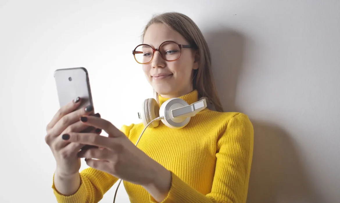 A content creator in a yellow jumper with headphones round her neck, holding a smartphone up in front of a white wall