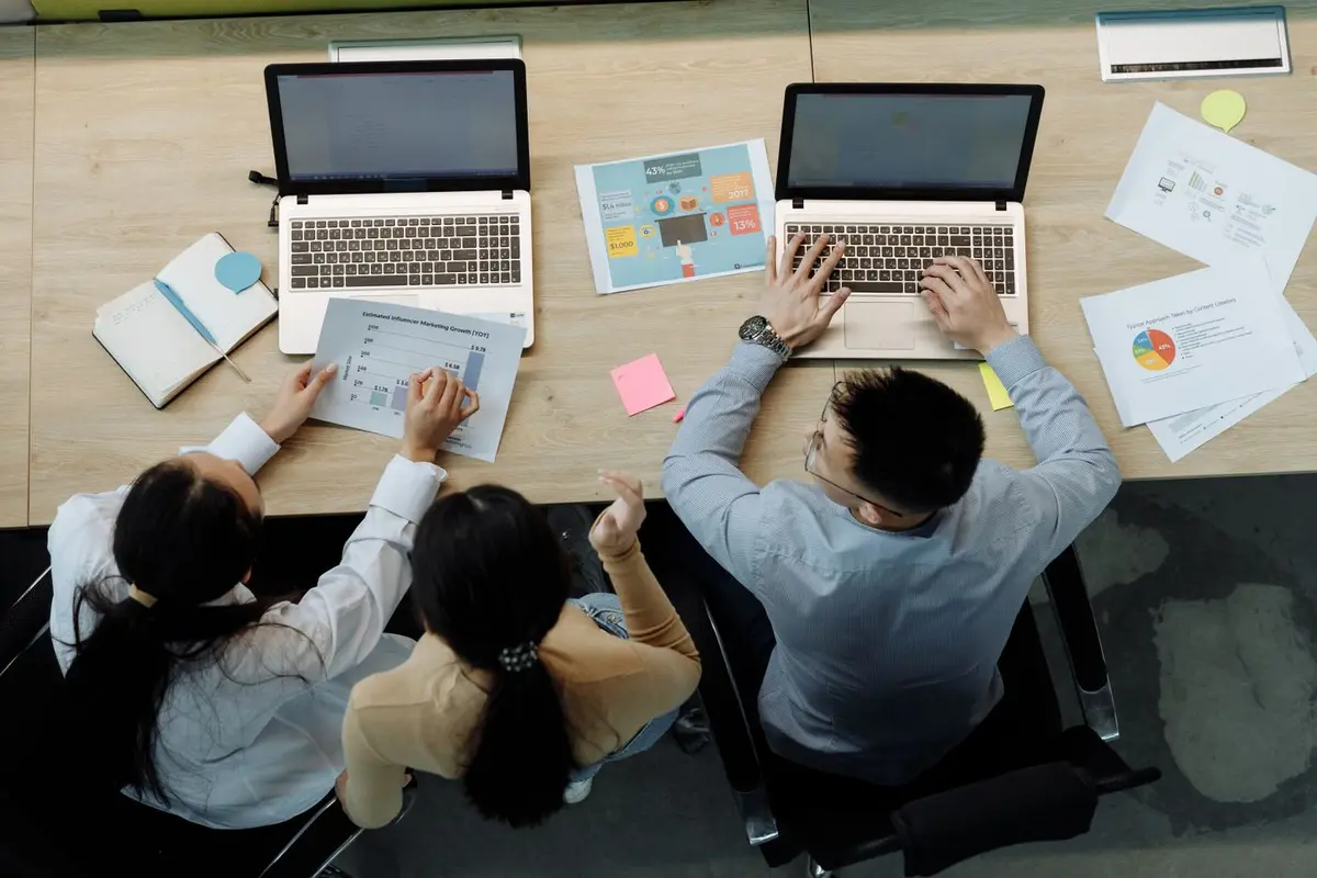 Three colleagues seen from directly above at a shared desk, reading printed charts and working on two laptops — the monthly read-out a local seo audit sets its baseline for.